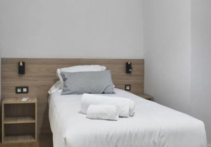 Habitación de hotel pequeña con una cama individual, ropa de cama blanca, almohada gris y cabecero de madera.
