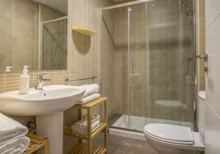 Pequeño baño de color beige con lavabo, ducha de cristal, inodoro y estantes de almacenamiento de mimbre.