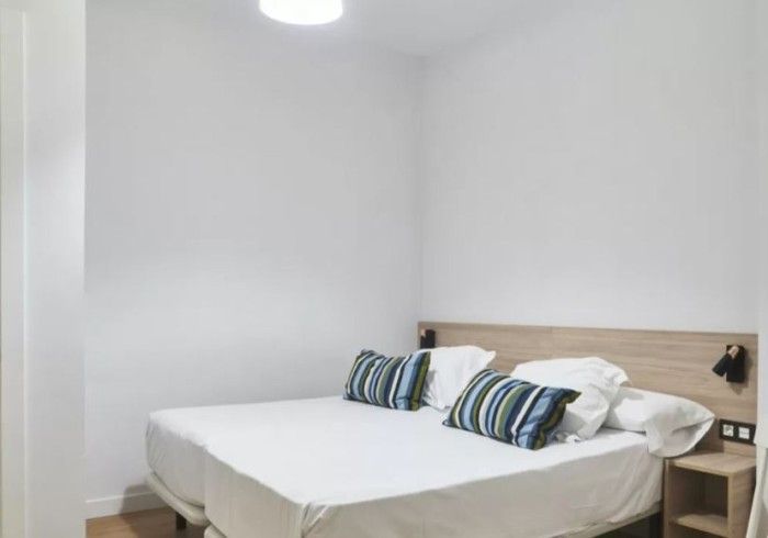 Habitación pequeña de color blanco con una cama, almohadas a rayas y una mesita de noche bajo una lámpara de techo.