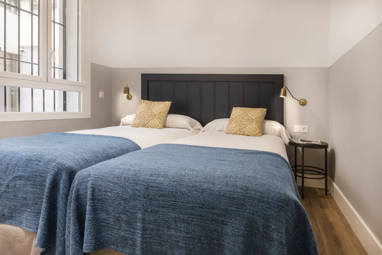 Habitación luminosa con dos camas con fundas azules, paredes beige y una ventana que deja entrar la luz natural.