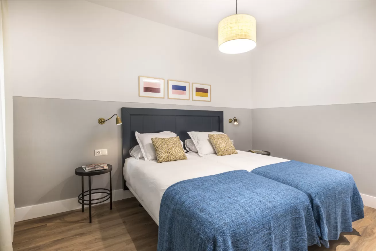 Habitación luminosa con colcha azul, cabecero gris, mesita de noche y lámpara de techo colgante.