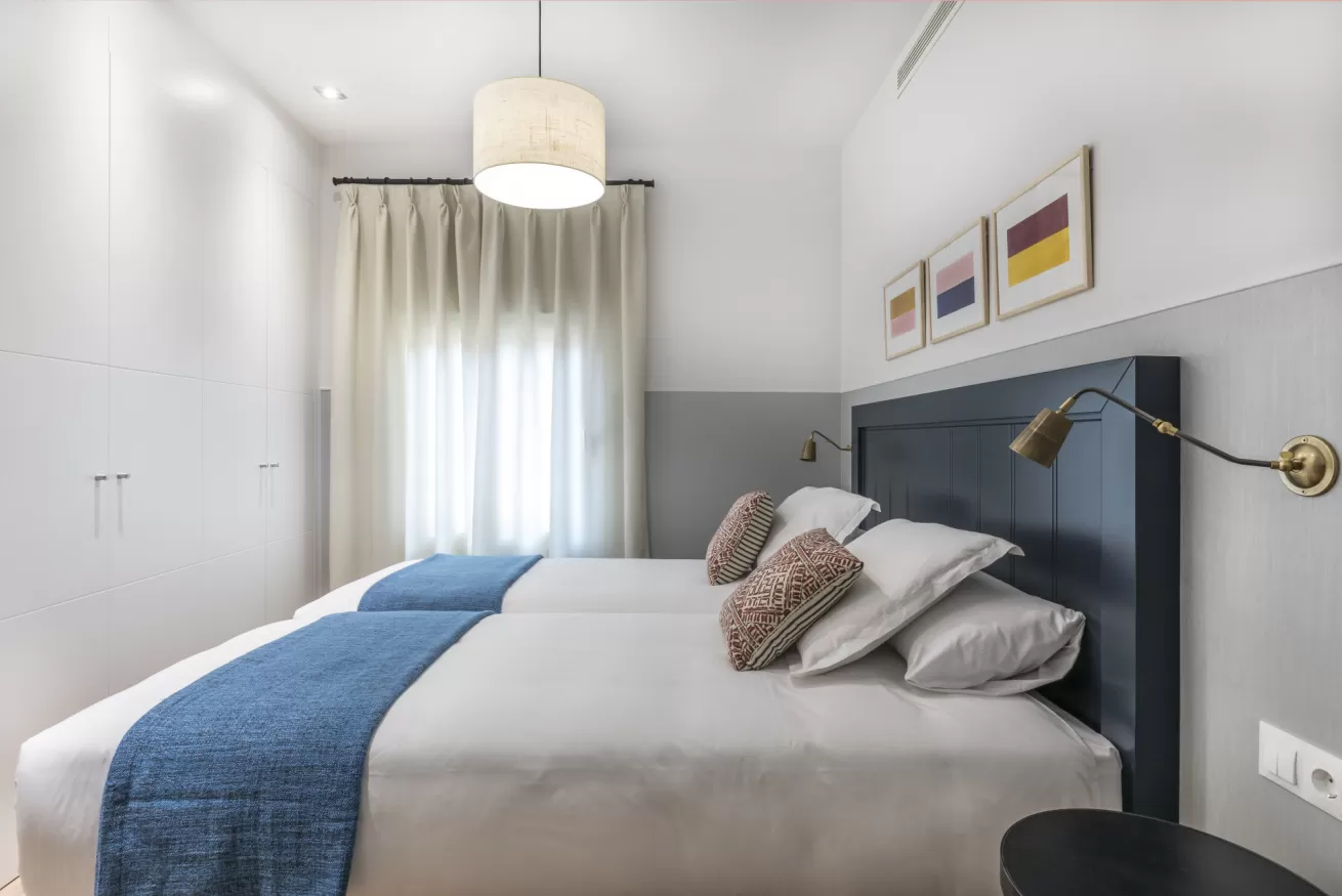 Dormitorio moderno con cama gris, manta azul, cortinas blancas y cuadro enmarcado en la pared.