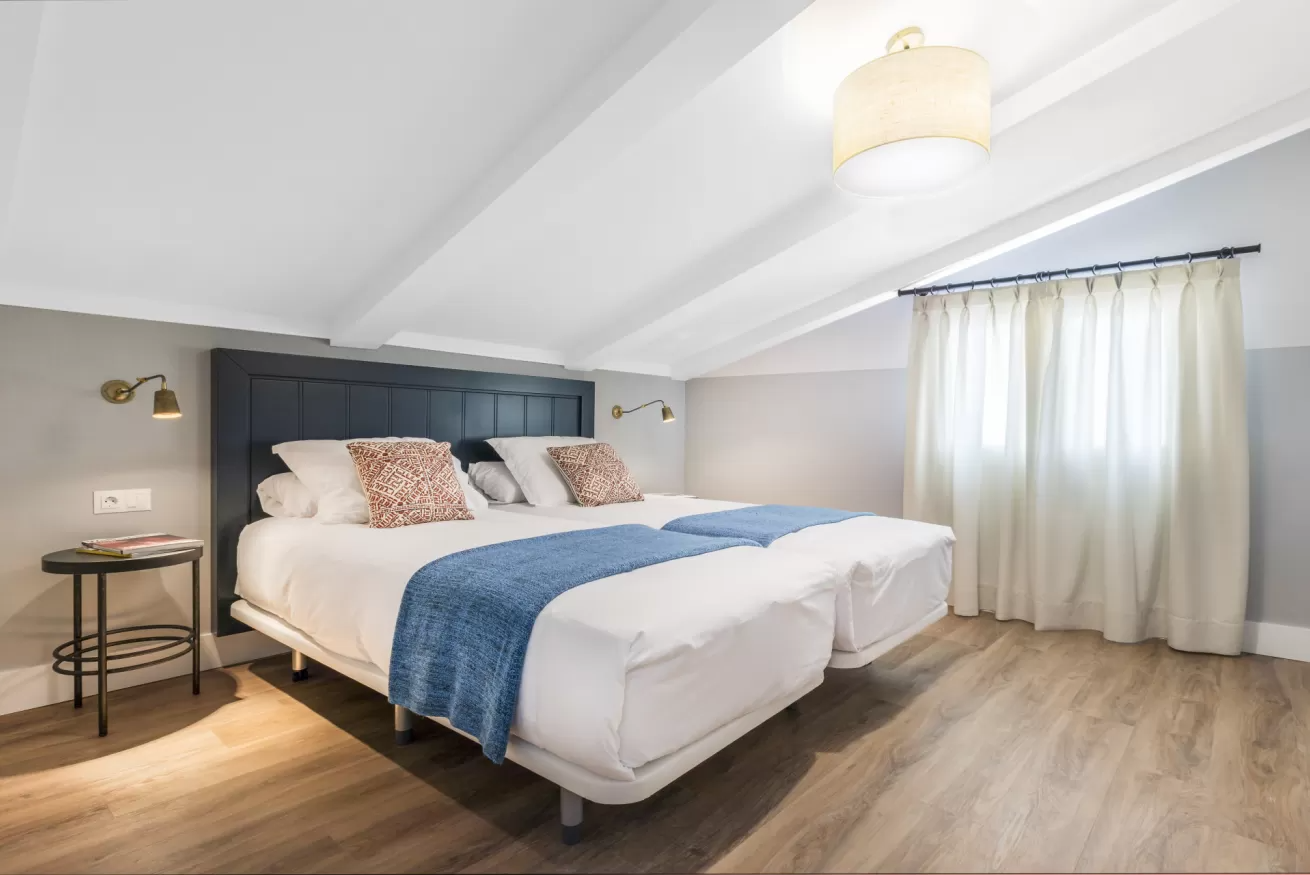 Habitación luminosa con una cama grande, una manta azul y cortinas transparentes junto a la ventana.