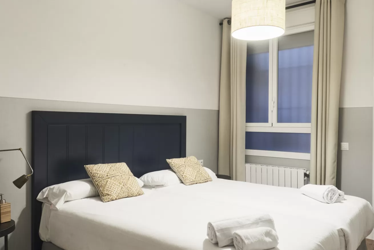 Habitación ordenada con una cama tapizada en color oscuro, ropa de cama blanca, cortinas beige y una ventana.