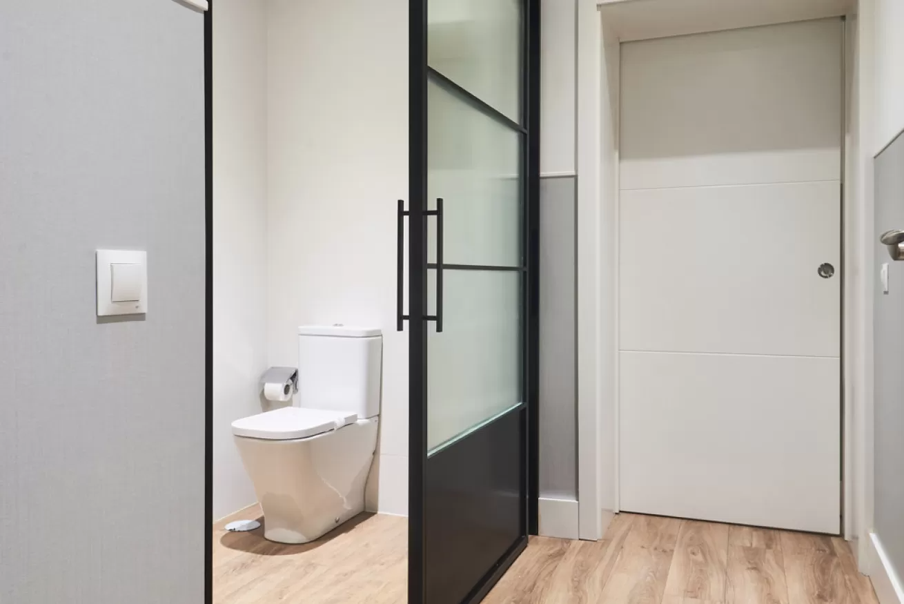 Pasillo con un pequeño baño, puerta corrediza de vidrio esmerilado y puerta de entrada blanca.