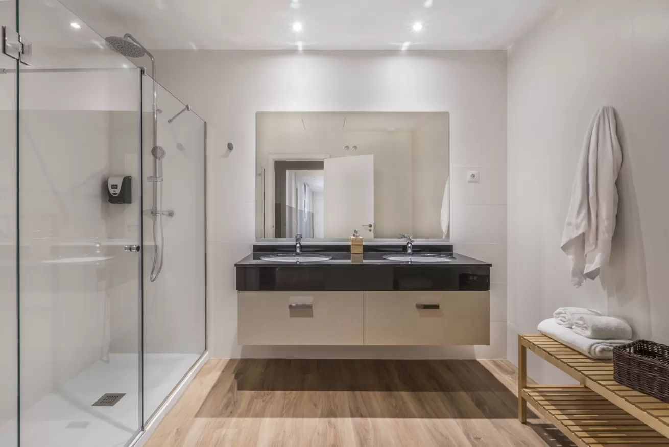 Baño moderno con ducha de cristal, lavabo doble, suelo de madera y toallero.