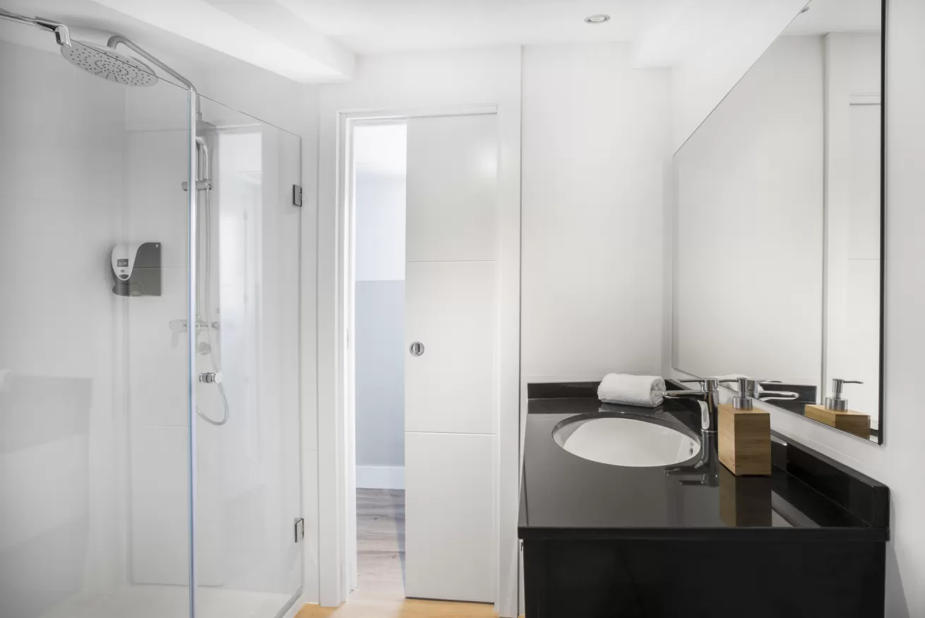 Baño moderno de color blanco con ducha, lavabo, espejo y puerta abierta que da a un pasillo alicatado.