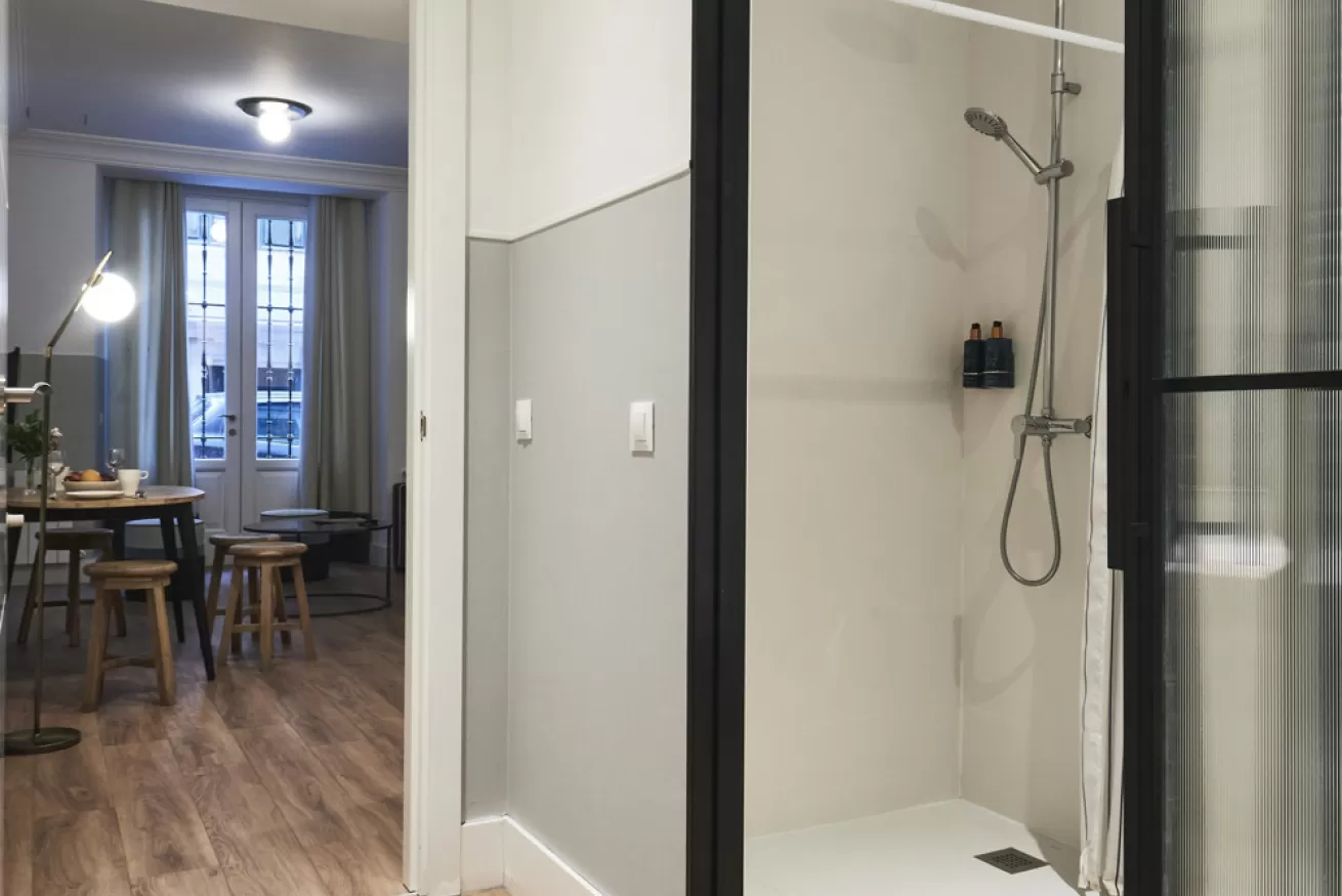 Pasillo de apartamento moderno con suelos de madera, zona de comedor y cabina de ducha de cristal a la derecha.