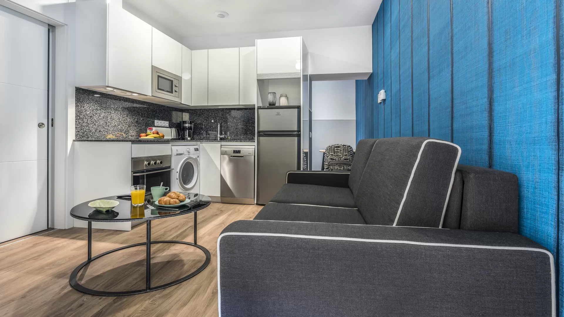 Apartamento tipo estudio moderno con sofá gris, pared decorativa azul y área de cocina compacta.