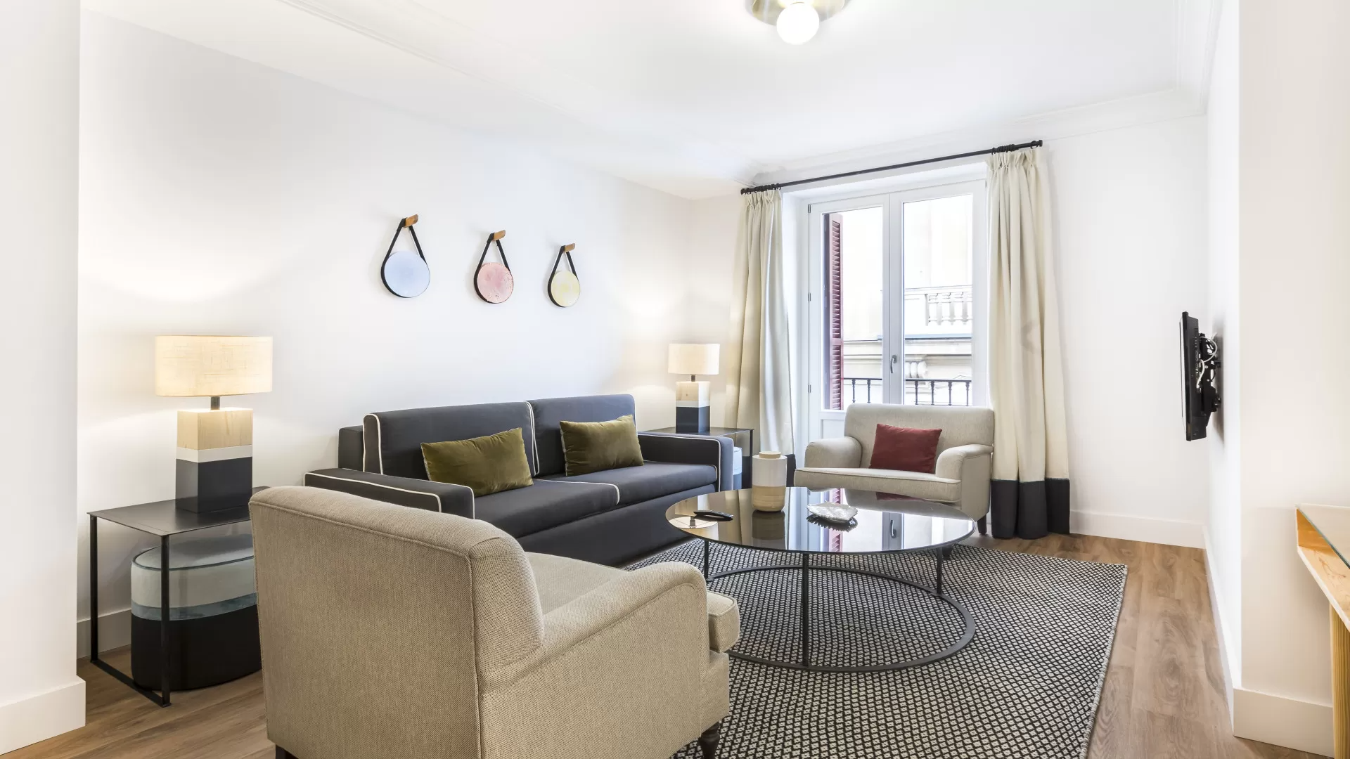 Sala de estar moderna y luminosa con sofá gris, sillón, mesa de centro, alfombra estampada y grandes cortinas.