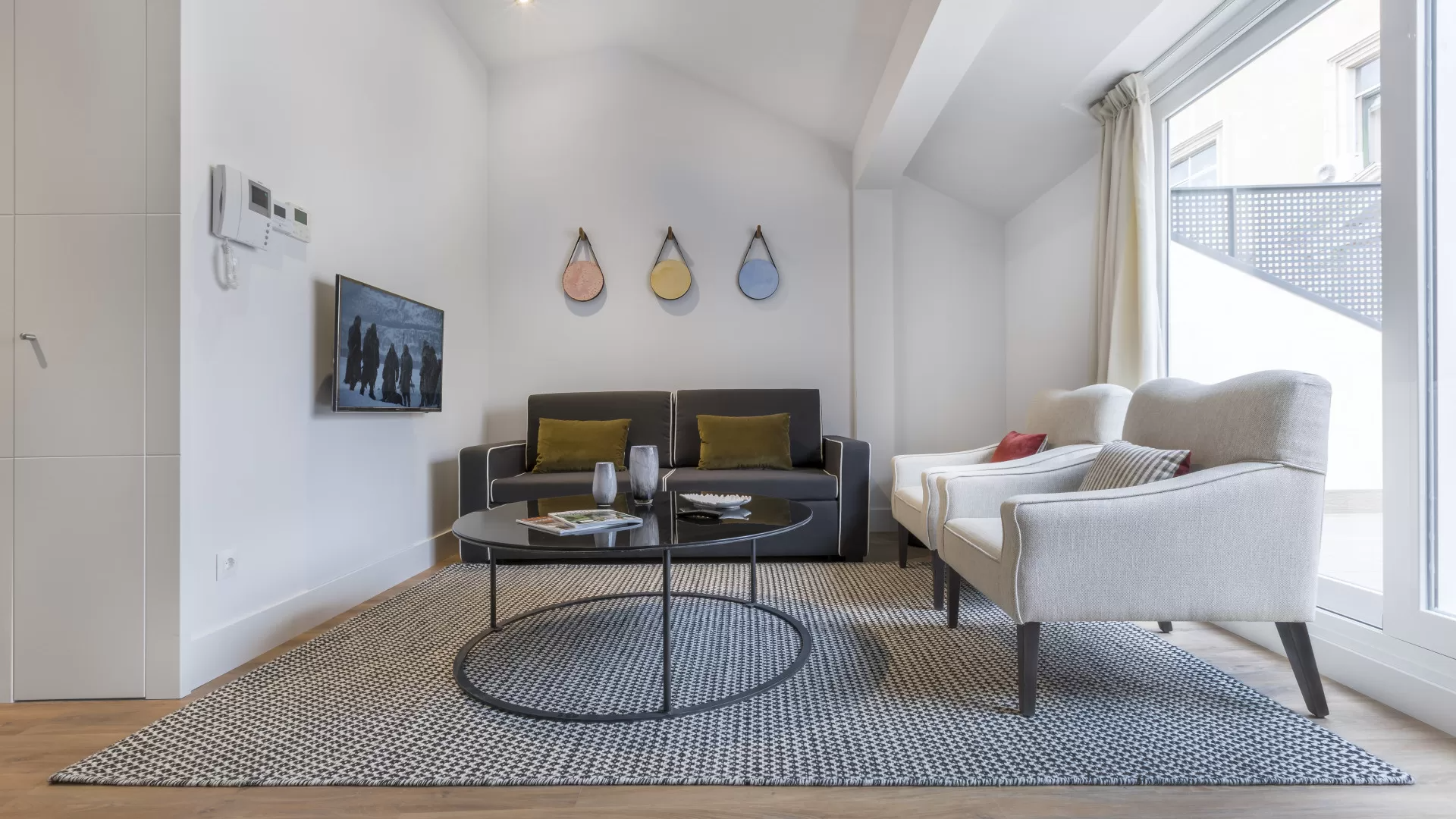 Luminoso salón moderno con sofá gris, silla blanca, alfombra estampada y grandes ventanales.