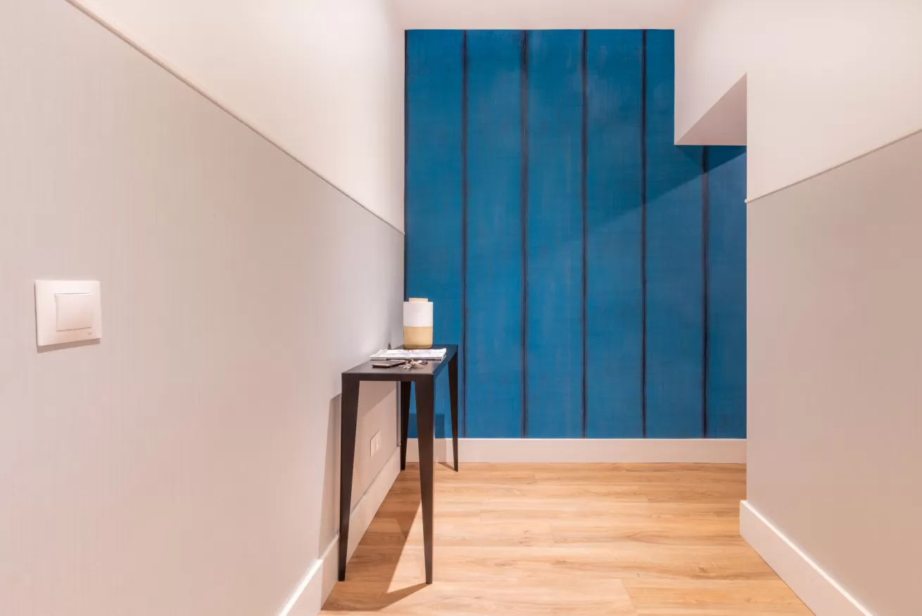 Pasillo estrecho con suelo de madera clara, paredes grises y una puerta con paneles azules al final.