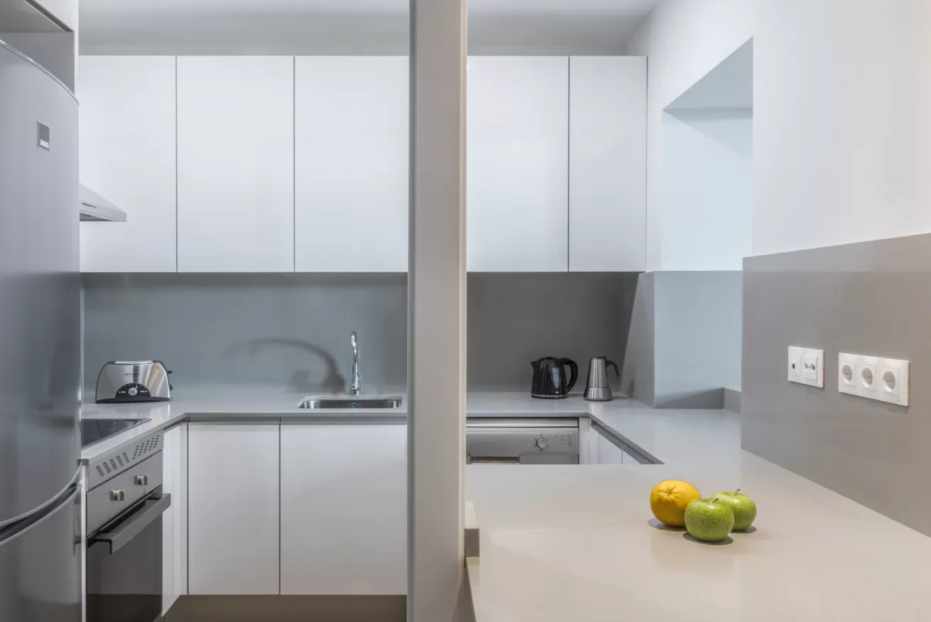 Cocina minimalista blanca con detalles en gris, fregadero, fogones, armarios y encimera con fruta y hervidor de agua.