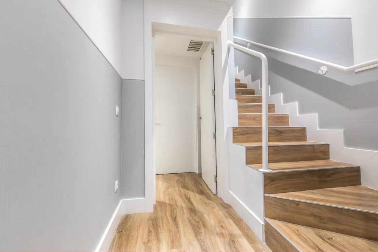 Pasillo luminoso con escaleras de madera, paredes grises y molduras blancas en un interior moderno.