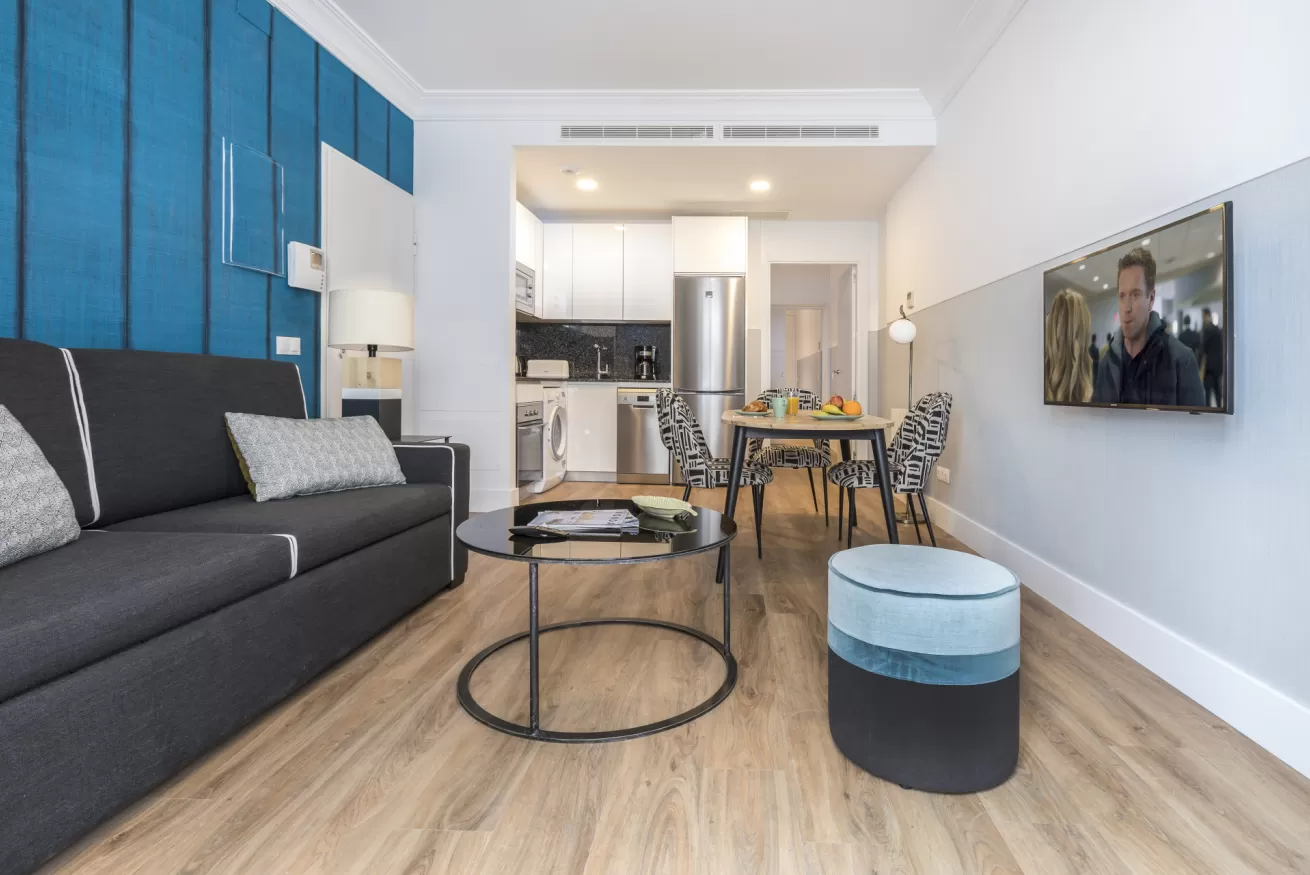 Sala de estar moderna con sofá oscuro, pared decorativa azul, mesa de centro redonda y zona de comedor más allá.
