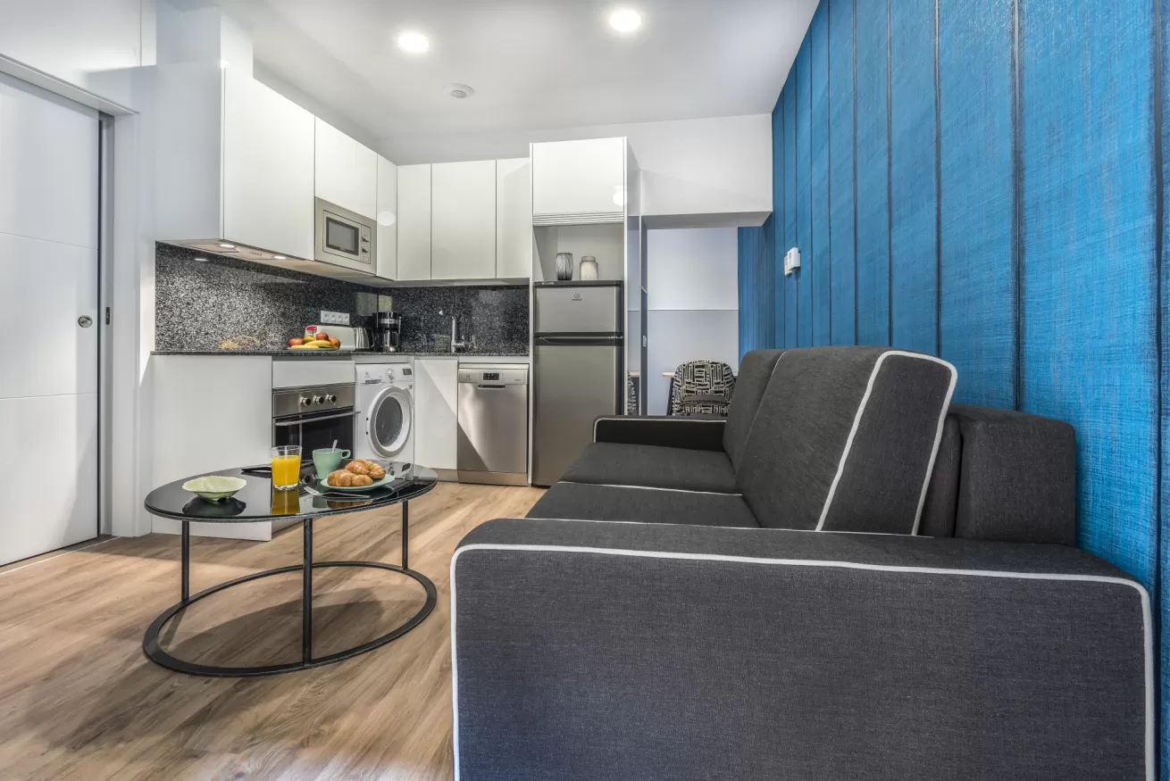 Apartamento tipo estudio moderno con sofá gris, mesa redonda de cristal, cocina blanca y pared decorativa azul.