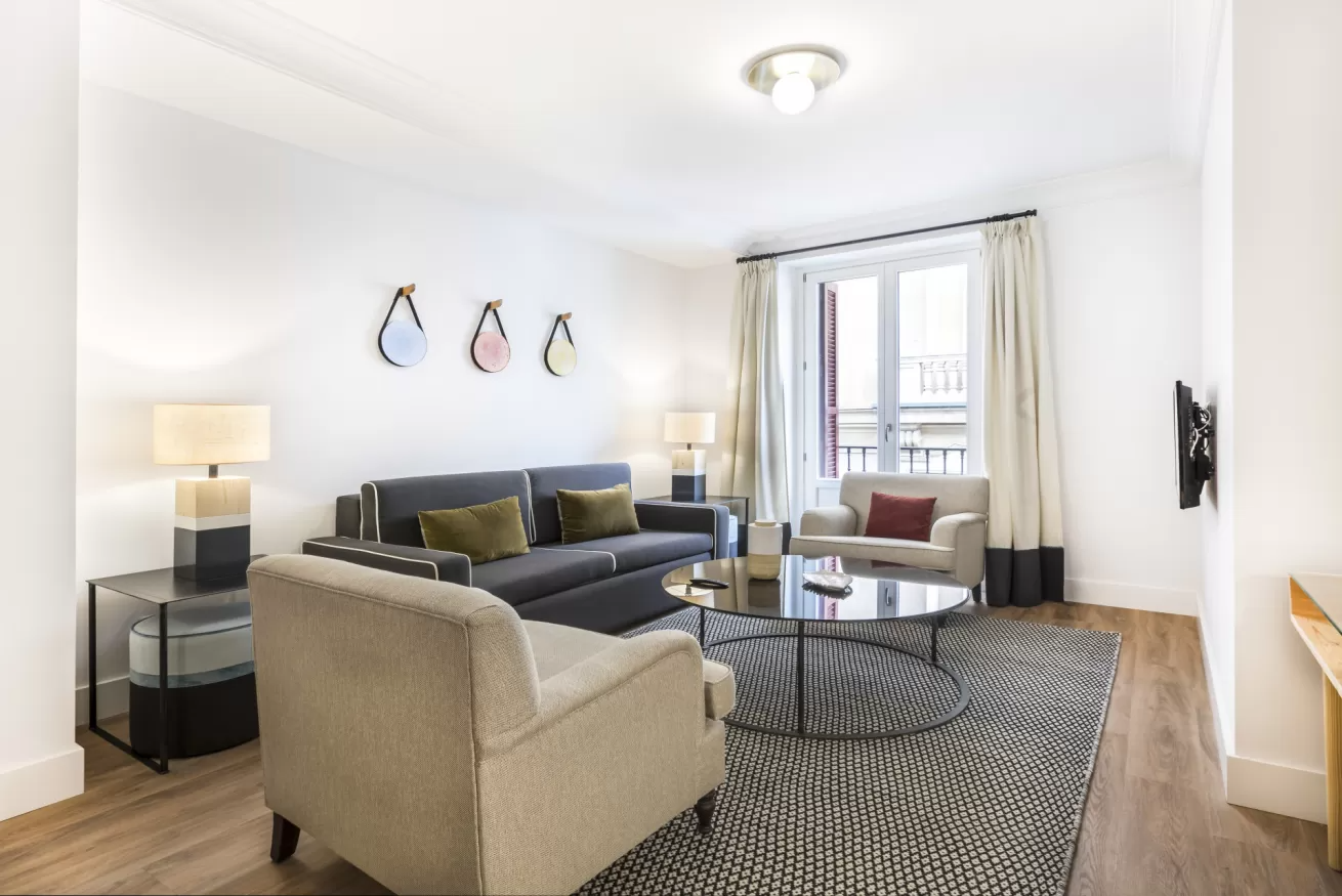 Sala de estar luminosa con sofá modular gris, silla beige, mesa de centro redonda, lámparas y una ventana con cortinas.