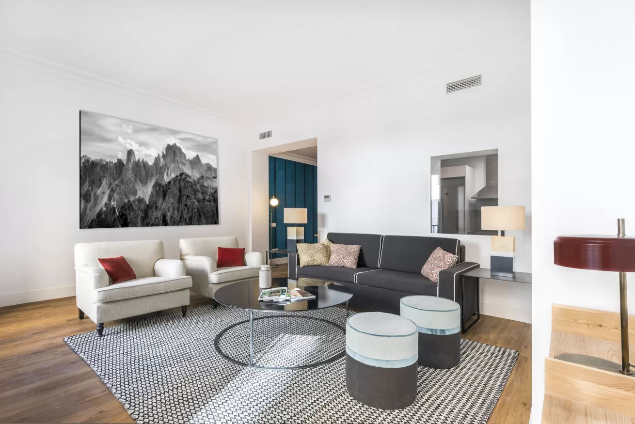 Sala de estar moderna con paredes blancas, sofá gris, sillones, alfombra estampada y un gran cuadro de montaña.