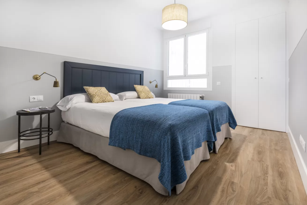 Dormitorio moderno con cama tapizada en gris, manta azul, suelo de madera y ventana luminosa.