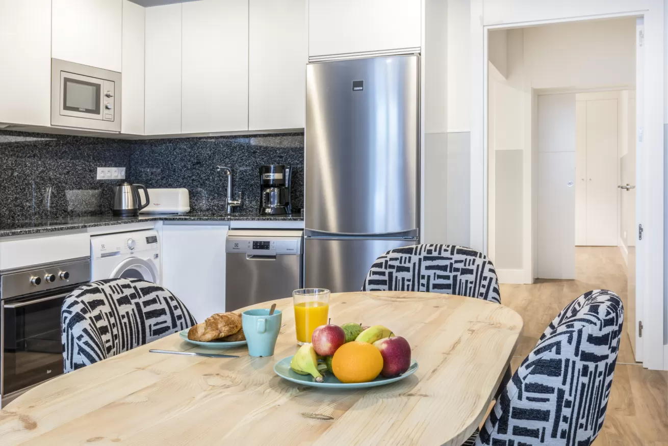 Cocina moderna con electrodomésticos de acero inoxidable y mesa de comedor con frutero y tazas.