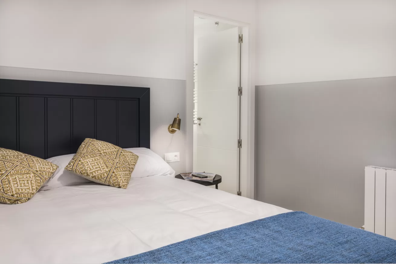 Habitación luminosa con cama blanca, cabecero negro, cojines estampados, manta azul y puerta abierta contra paredes grises.