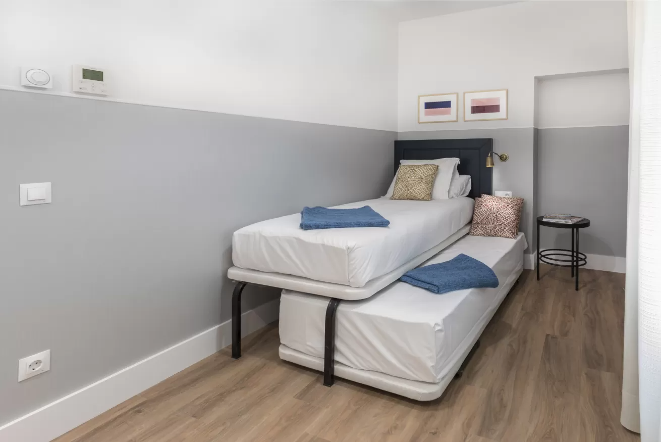 Habitación pequeña con dos camas individuales, paredes grises, suelo de madera y una mesita de noche estrecha.