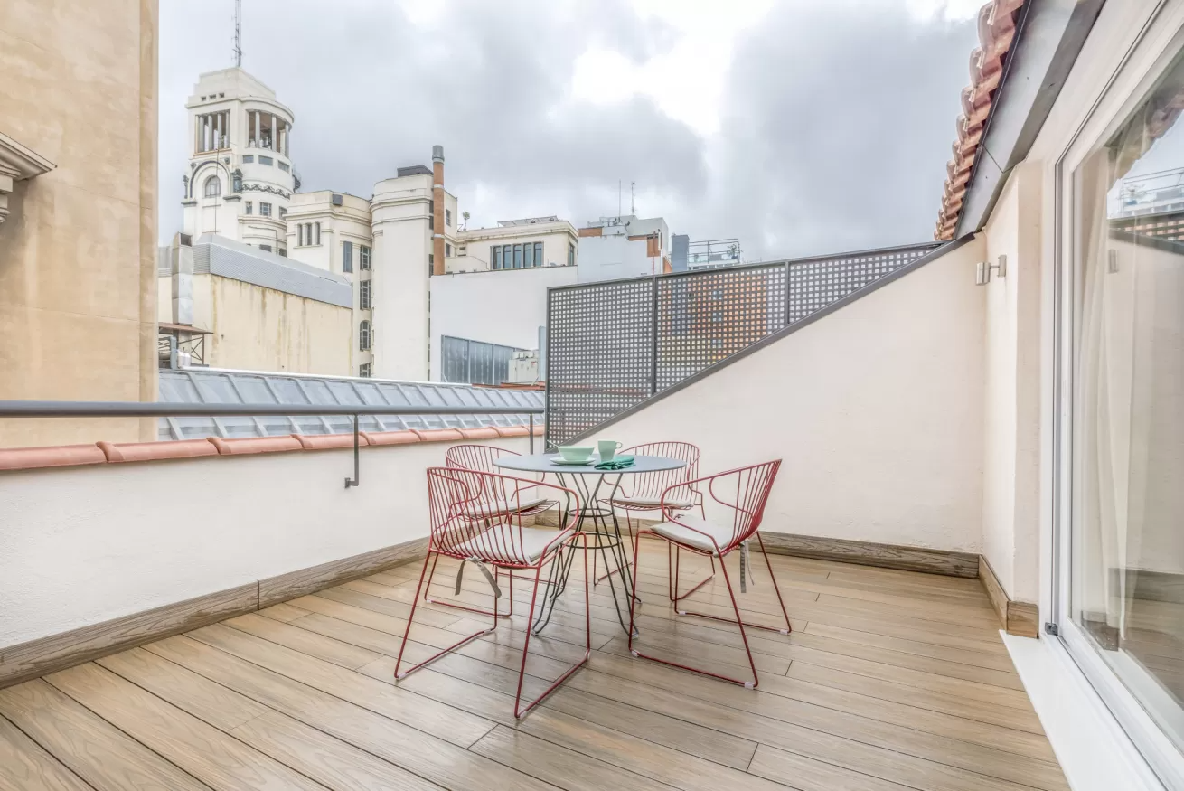Terraza en la azotea con sillas y mesa de alambre, tarima de madera clara, edificios de la ciudad al fondo.