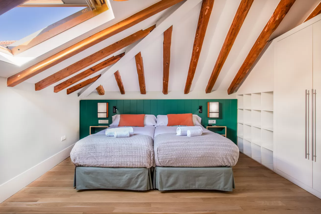 Luminoso dormitorio abuhardillado con dos camas individuales, cabecero verde, vigas de madera a la vista y ventanas de techo.