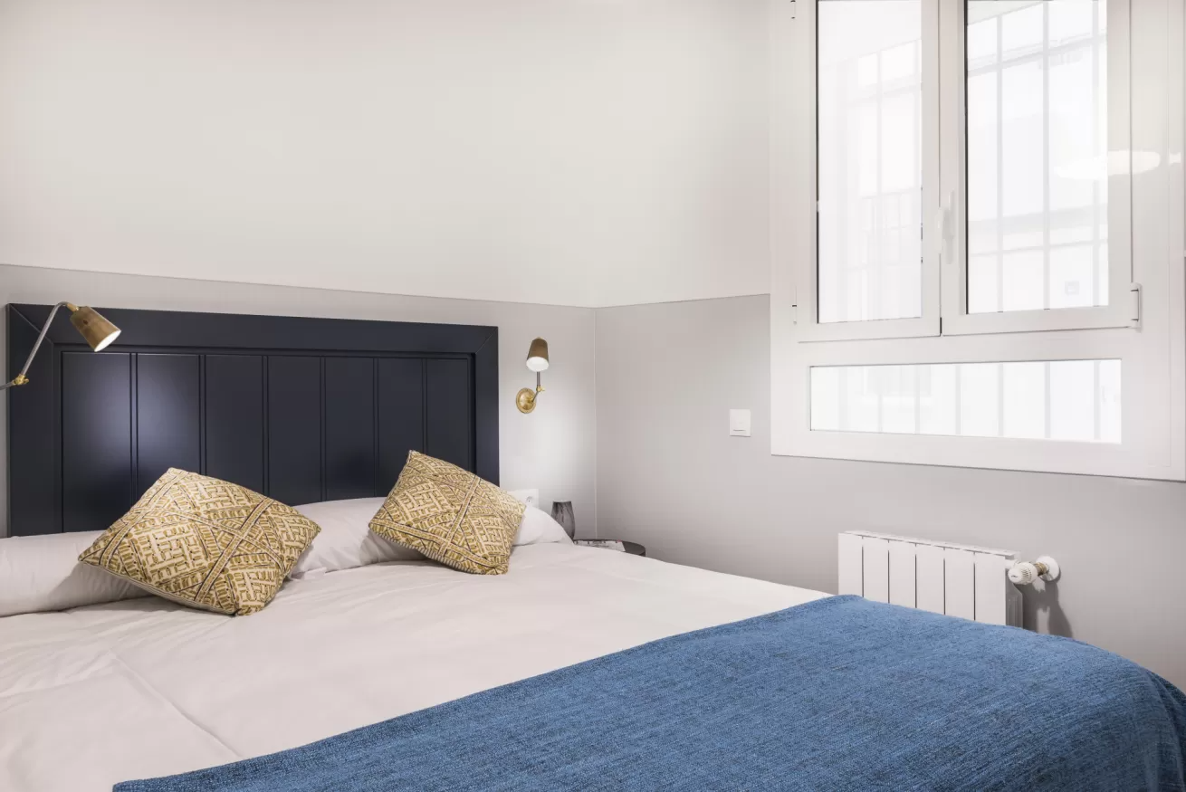 Habitación luminosa con cabecero oscuro, paredes blancas, colcha azul y una ventana sobre un radiador blanco.