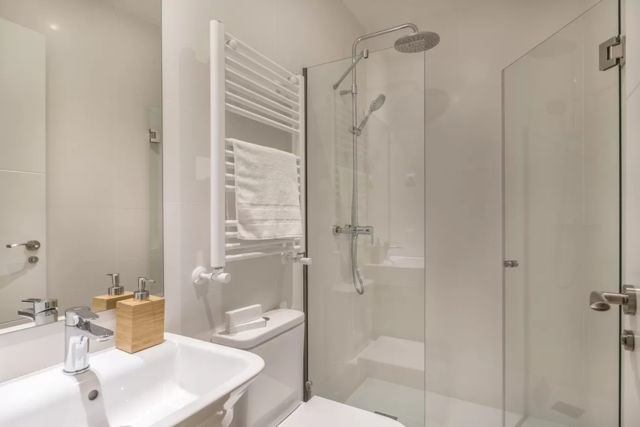 Baño moderno blanco con lavabo, radiador toallero y cabina de ducha de cristal.