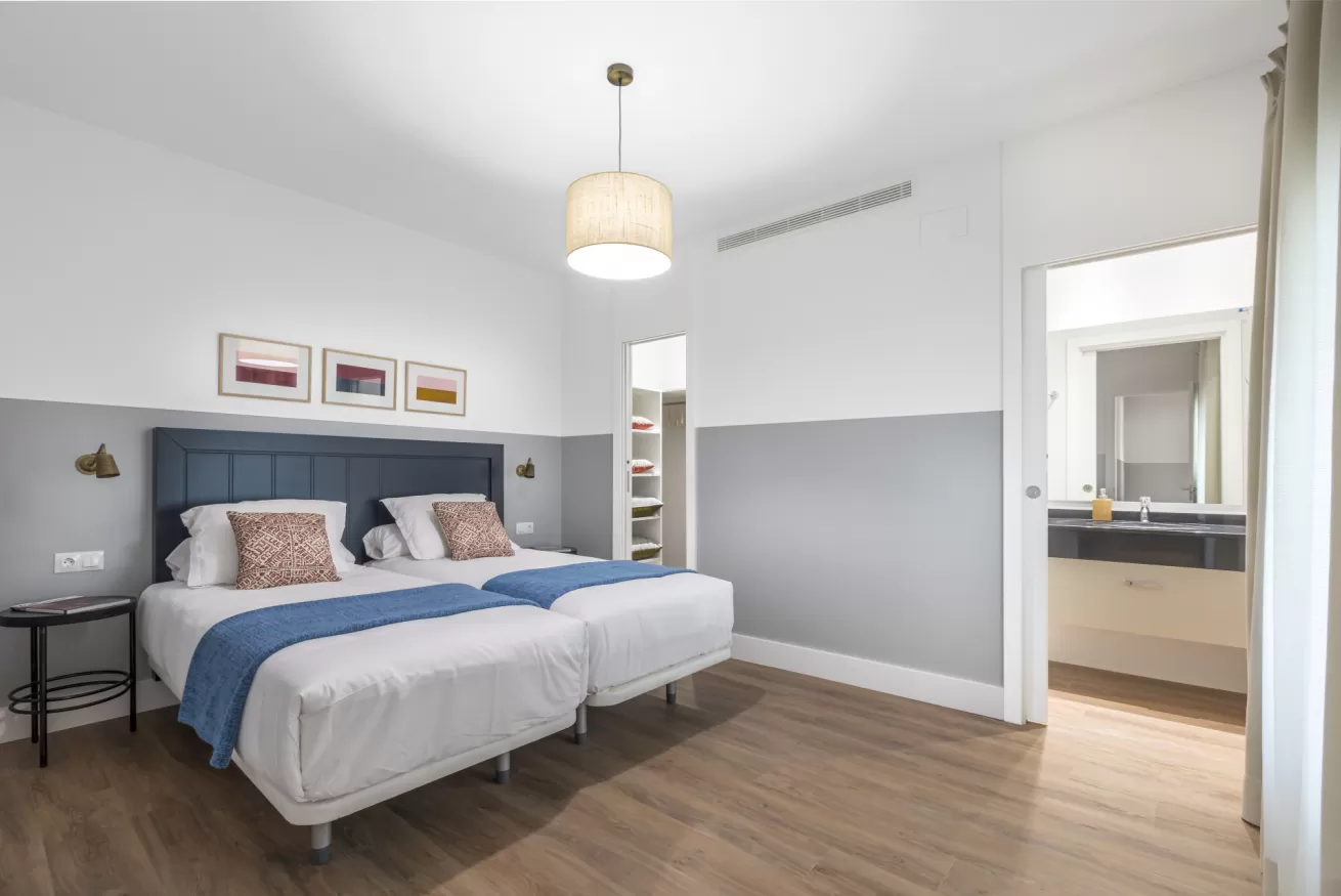 Habitación luminosa con cama tapizada en gris, detalles en azul, suelo de madera y puerta abierta que da al baño.