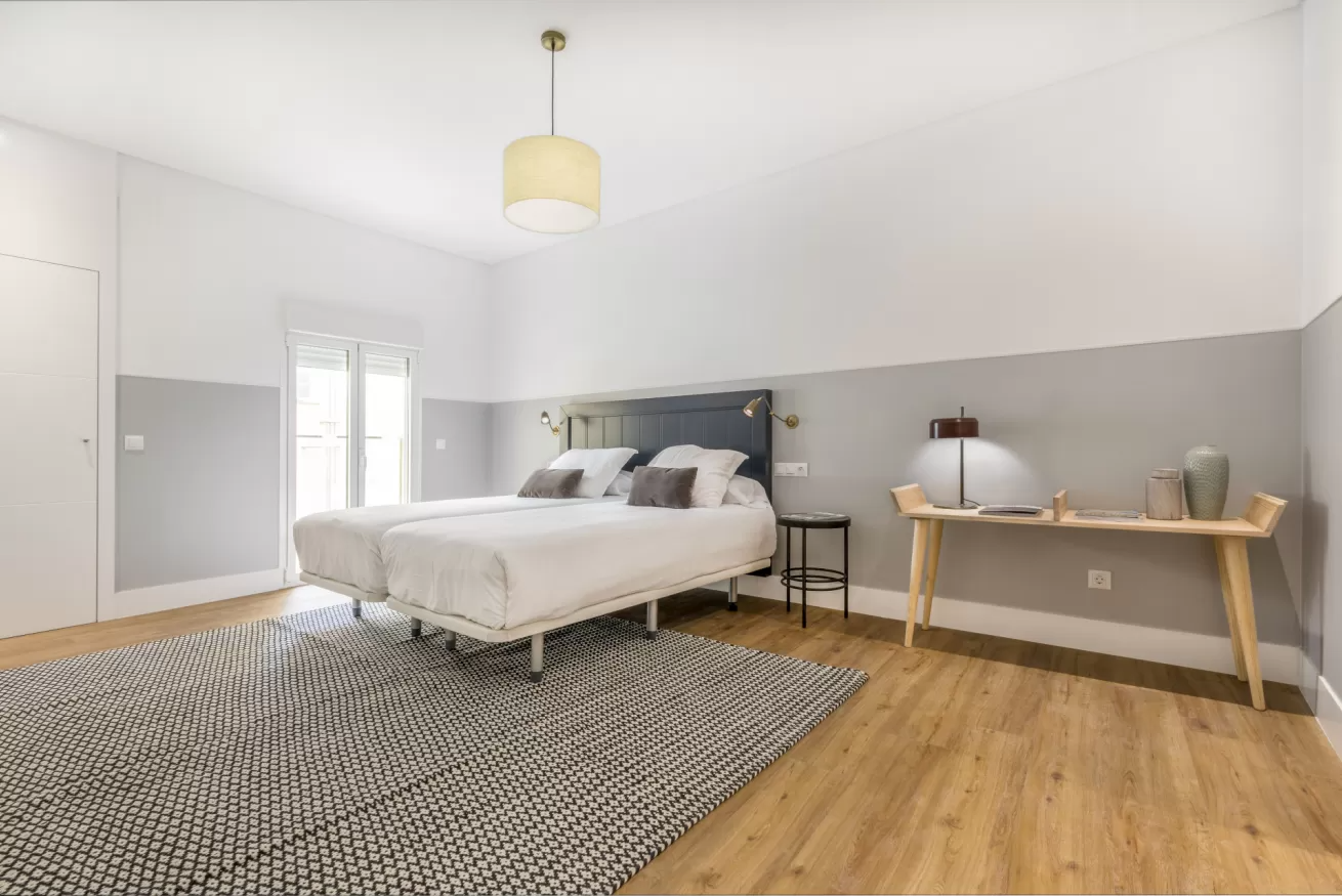 Habitación luminosa y minimalista con cama, alfombra estampada, suelo de madera y escritorio junto a la ventana.