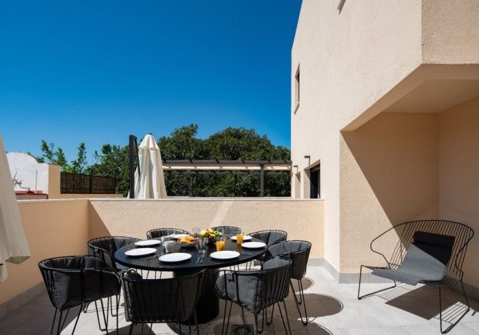 Patio exterior con mesa de comedor redonda negra, sillas de mimbre y un edificio beige bajo un cielo azul brillante.