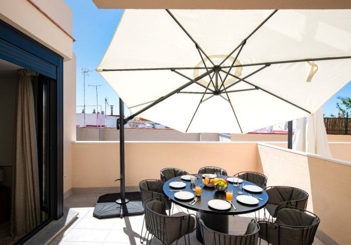 Mesa de comedor exterior en el patio bajo una gran sombrilla blanca con platos y vasos junto a un balcón con vistas
