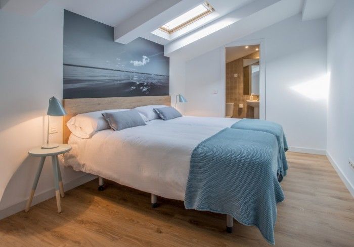 Habitación luminosa con tragaluces, cama gris, manta azul y lámpara de noche.