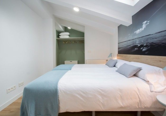 Habitación luminosa con cama blanca, manta azul claro y cuadro con motivos marinos bajo un techo inclinado.