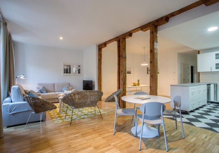 Amplio salón-comedor diáfano con sofá azul, silla de mimbre, suelos de madera y cocina contigua.