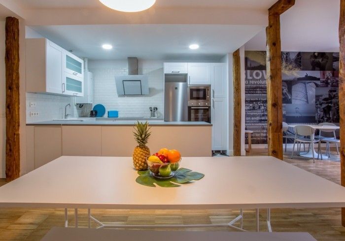 Cocina y comedor modernos con mesa blanca, centro de mesa con frutas y vigas de madera a la vista.