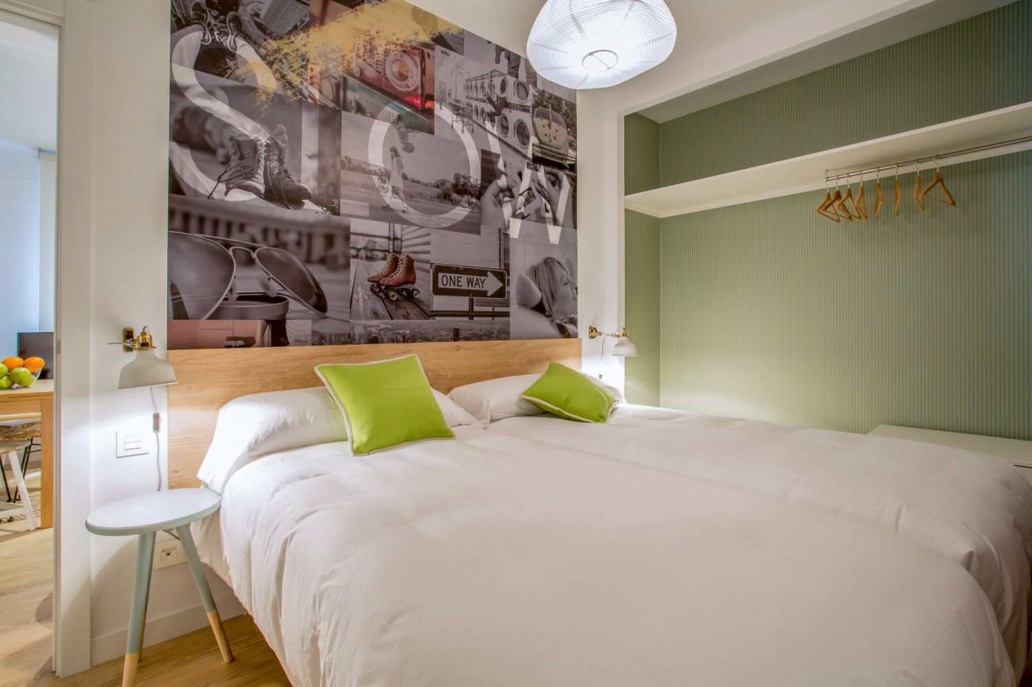 Habitación luminosa con cama blanca, almohadas verde lima, mural en la pared y armario empotrado abierto.