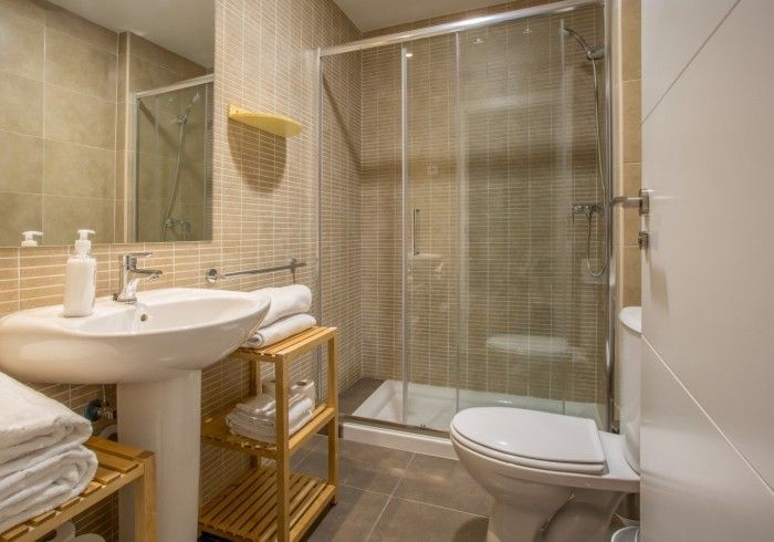 Pequeño baño de color beige con lavabo, ducha, inodoro y estantes de mimbre.