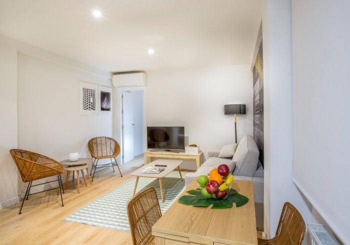 Luminoso salón con sofá blanco, sillas de mimbre, televisor pequeño, alfombra a rayas y frutero sobre una mesa de madera.