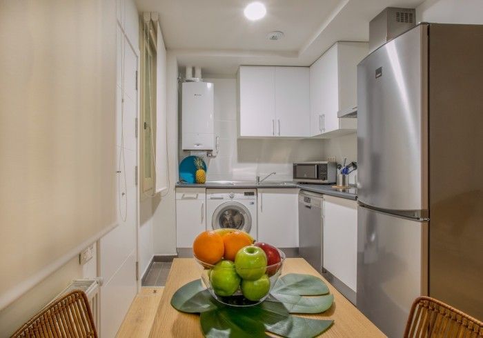 Cocina luminosa y compacta con armarios blancos, nevera de acero inoxidable y un frutero sobre una mesita.