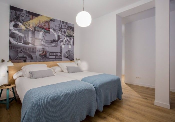 Habitación luminosa con dos camas con fundas azules, un mural gris en la pared, suelo de madera y una lámpara blanca .