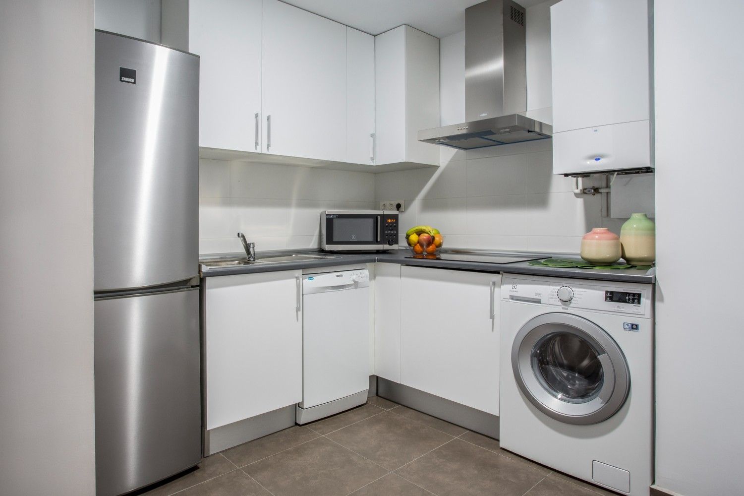 Cocina compacta blanca con nevera, lavadora, microondas, fregadero y frutero de acero inoxidable.