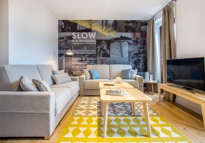 Sala de estar moderna con sofá gris, sillón blanco, mesa de centro de madera, televisor y alfombra geométrica amarilla.