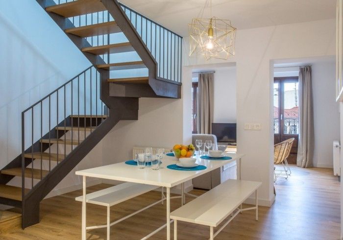 Comedor moderno con escalera, mesa blanca y habitación contigua luminosa.