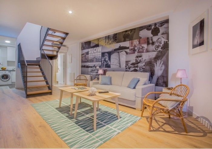 Sala de estar moderna con sofá gris, mesas de centro de madera, alfombra a rayas, silla de mimbre y escalera.