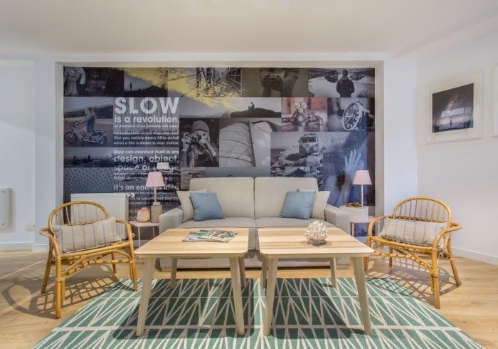 Salón luminoso con sillas de mimbre, mesa de madera, alfombra estampada y un mural en la pared.