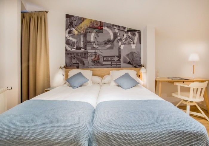 Habitación luminosa con dos camas individuales, ropa de cama azul, cuadros enmarcados en la pared y una pequeña lámpara.