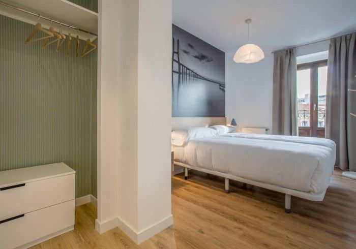 Dormitorio moderno con cama blanca, suelo de madera, armario a rayas verdes y una gran ventana.